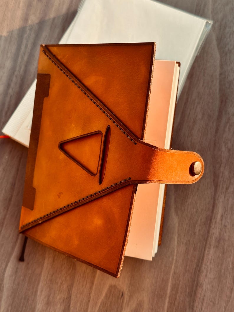 Pu&ograve; includere: Un diario in pelle color cuoio con un design a intaglio triangolare sulla copertina anteriore. La copertina &egrave; fissata con un cinturino in pelle abbinato che include una chiusura a bottone automatico in metallo. Il diario contiene pagine bianche.
