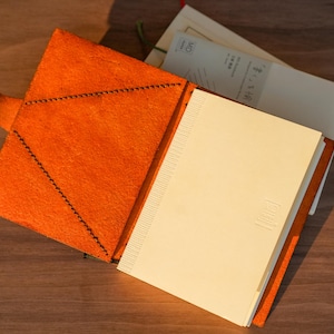 Pu&ograve; includere: Una copertina per taccuino in pelle arancione aperta con chiusura a bottone automatico e cuciture scure visibili. La copertina contiene un taccuino in carta color crema con dorso testurizzato e un piccolo logo in rilievo sulla prima pagina.