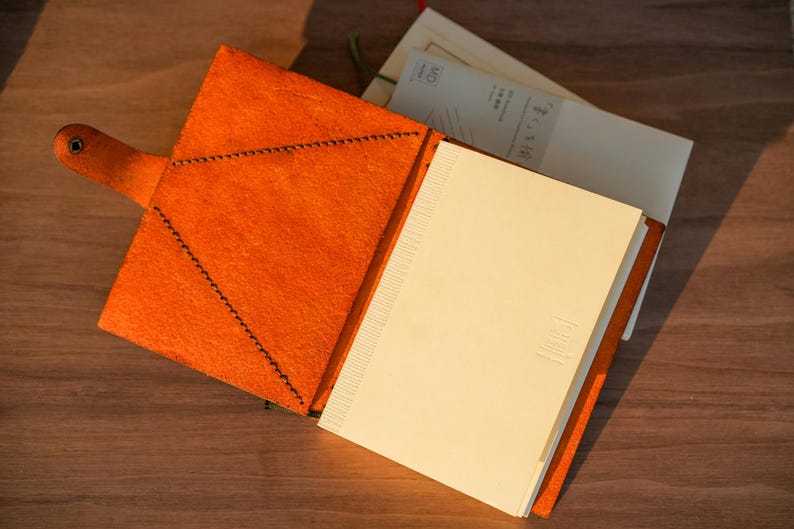 Pu&ograve; includere: Una copertina per taccuino in pelle arancione aperta con chiusura a bottone automatico e cuciture scure visibili. La copertina contiene un taccuino in carta color crema con dorso testurizzato e un piccolo logo in rilievo sulla prima pagina.