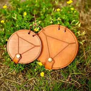May include: Two round, wooden coasters with a geometric design. The coasters are a warm brown color with a lighter brown triangle design in the center. Each coaster has a leather strap and a metal button. The coasters are sitting on grass with yellow flowers.