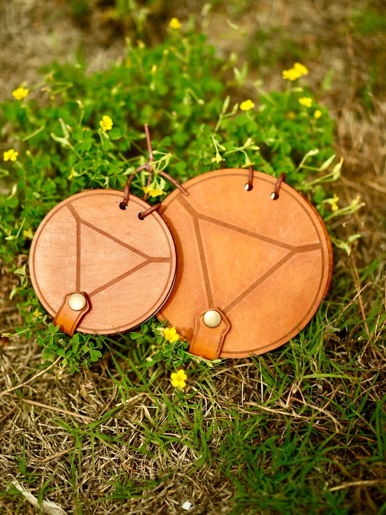 May include: Two round, wooden coasters with a geometric design. The coasters are a warm brown color with a lighter brown triangle design in the center. Each coaster has a leather strap and a metal button. The coasters are sitting on grass with yellow flowers.