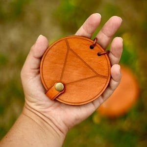 May include: A round, orange-brown leather coaster with a geometric design. The coaster has a triangle pattern etched into the surface and a small leather strap with a brass button. Two small holes are at the top with a brown string.
