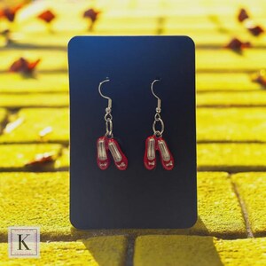 May include: A pair of silver dangle earrings featuring red ballet slipper charms. The earrings are displayed on a black card against a yellow background with scattered leaves. The ballet slippers have white accents.