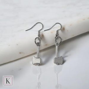 May include: A pair of silver-colored axe-shaped earrings. Each earring features a small axe pendant suspended from a hook. The axes have a detailed design, and the earrings are displayed against a white background.