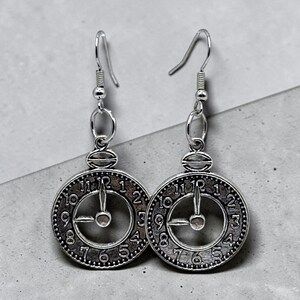 May include: Silver-toned clock face earrings with a hook closure. Each earring features a circular clock design with Roman numerals and clock hands. The earrings are suspended from a silver hook.