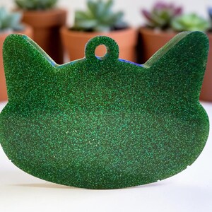 May include: A green glitter cat-shaped pet tag with a small hole at the top for attaching to a collar. The tag is in front of several potted succulents.