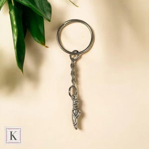 May include: Silver-toned keychain featuring a ballet slipper charm. The charm is detailed with delicate lines and textures, hanging from a chain and a large ring. The background is a soft, neutral color, with green leaves in the upper left corner.