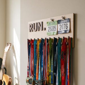 Puede incluir: Un tablero de exhibición de medallas de madera con la palabra "RUN" en letras de madera. El tablero contiene numerosas medallas de carrera coloridas y dos dorsales con los números 2605 y 2076. Las medallas tienen cintas de varios colores.