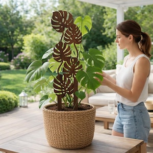 May include: A brown metal plant trellis shaped like Monstera leaves supports a large potted plant with green leaves. The plant is in a woven basket planter. A person is tending to the plant on a wooden deck.