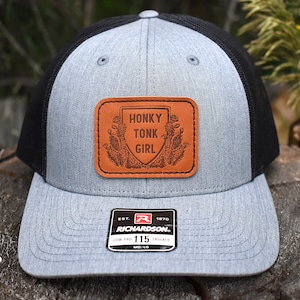 May include: A gray and black trucker hat with a brown leather patch that says "Honky Tonk Girl". The patch has a floral design around the text. The hat is made by Richardson and has a size tag that says "115".