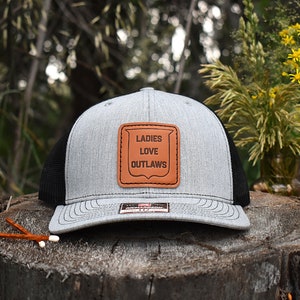 May include: A gray and black trucker hat with a brown leather patch that says "LADIES LOVE OUTLAWS".