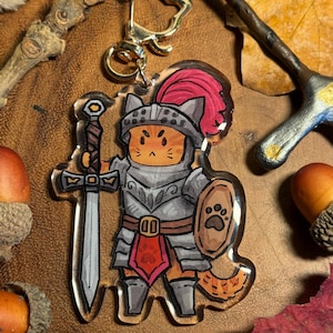 May include: A clear acrylic keyring depicting a cartoon cat in knight's armour, holding a sword and shield with a paw print. The cat has orange fur, a red plume, and is attached to a gold-coloured clasp.