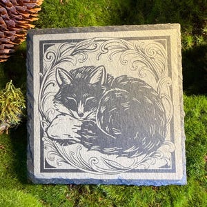 May include: A square slate coaster featuring a laser-engraved illustration of a curled-up sleeping fox. The fox is centred within a decorative border of swirling filigree patterns. The natural stone has a dark grey colour and a rough, textured edge.