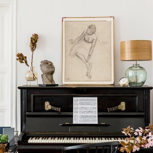 K&ouml;nnte beinhalten: Ein schwarzer Vintage-Klavier mit einem Notenblatt auf der Oberseite. Ein Ballerina-Zeichnung h&auml;ngt an der Wand &uuml;ber dem Klavier. Eine Lampe mit einem Holzschirm und einer Glasbasis steht auf dem Klavier. Eine Vase mit getrockneten Blumen und eine Kopfskulptur stehen ebenfalls auf dem Klavier.