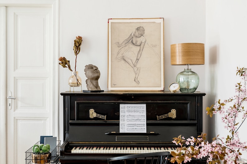 K&ouml;nnte beinhalten: Ein schwarzer Vintage-Klavier mit einem Notenblatt auf der Oberseite. Ein Ballerina-Zeichnung h&auml;ngt an der Wand &uuml;ber dem Klavier. Eine Lampe mit einem Holzschirm und einer Glasbasis steht auf dem Klavier. Eine Vase mit getrockneten Blumen und eine Kopfskulptur stehen ebenfalls auf dem Klavier.