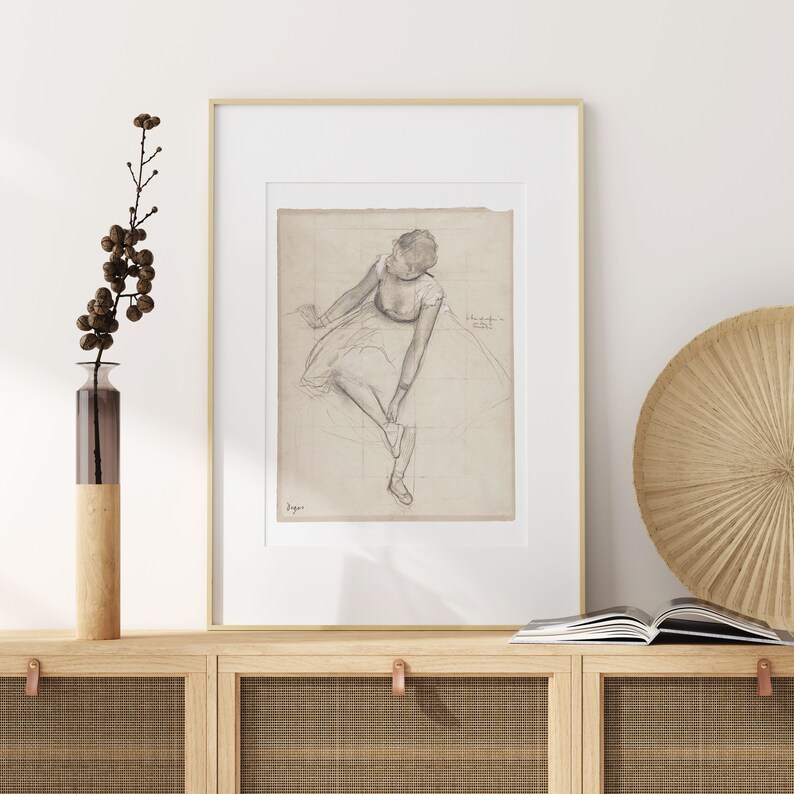 K&ouml;nnte beinhalten: Eine gerahmte Schwarzwei&szlig;-Bleistiftzeichnung einer Ballerina, die sitzt und ihren Schuh anpasst. Die Zeichnung ist auf hellbraunem Papier mit einem Gittermuster. Der Rahmen ist in schlichtem Gold.
