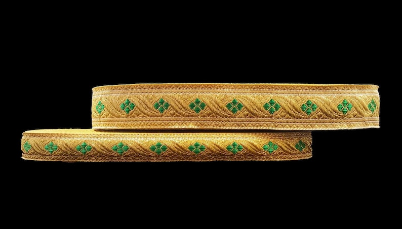 May include: Two gold-colored decorative ribbons with a repeating pattern of green floral designs. The ribbons are stacked on top of each other against a black background. The ribbons are likely used for embellishments.