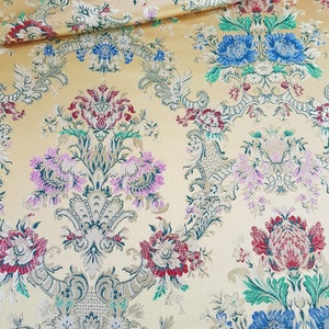 Könnte beinhalten: Ein goldfarbener Stoff mit einem floralen Muster. Das Muster zeigt rosa, rote, blaue und grüne Blumen mit goldenen Akzenten. Der Stoff hat eine Damast-ähnliche Textur.