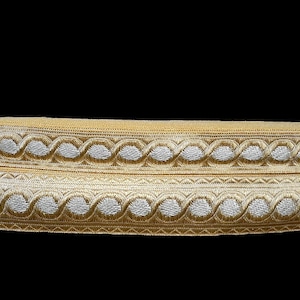 May include: Two rolls of decorative trim with a gold and silver design. The trim features a repeating pattern of interlocking circles and is likely used for embellishing clothing or crafts. The background is black.
