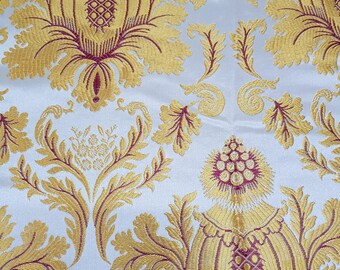Pode incluir: Tecido branco com um padrão repetido de desenhos florais dourados e bordô. O tecido tem uma textura em relevo.