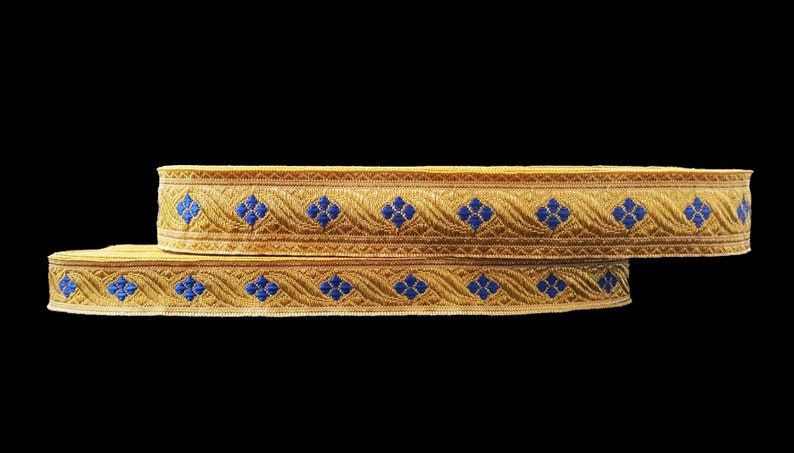May include: Two rolls of gold-colored decorative trim with a repeating pattern of blue floral shapes. The trim has a woven texture and is set against a black background, highlighting the intricate design.