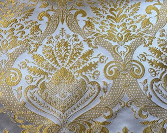 Pode incluir: Um tecido com um padrão de damasco repetitivo em fio dourado metálico tecido sobre um fundo liso branco sujo. O design consiste em intrincados motivos florais e de volutas com detalhes texturizados e formas simétricas.