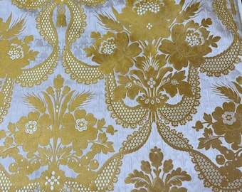 Puede incluir: Una tela con un patrón de damasco repetitivo en oro metálico y blanco. El diseño consiste en grandes motivos florales, intrincados trabajos de celosía y elementos de follaje estilizados tejidos en el textil, creando una apariencia texturizada.