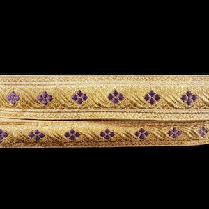 May include: Two gold-colored decorative ribbons with a repeating pattern of purple floral designs. The ribbons are layered on top of each other against a black background. The ribbons have a textured appearance.