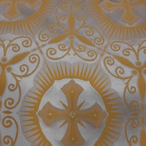 Può includere: Un tessuto bianco con un motivo dorato di croci e volute. Il tessuto ha un aspetto testurizzato e tessuto.