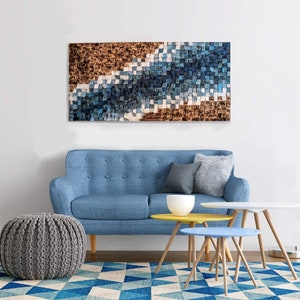May include: A blue and brown abstract wall art piece hangs above a blue sofa with two throw pillows. The sofa is in a living room setting with a grey knitted ottoman and two wooden coffee tables.