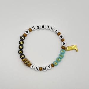 May include: A beaded bracelet featuring white, black, and turquoise letter beads spelling out THERE'S A SNAKE IN MY BOOT. The design includes brown spacer beads, a gold-toned snake charm, and a gold-toned cowboy boot charm attached to the side.
