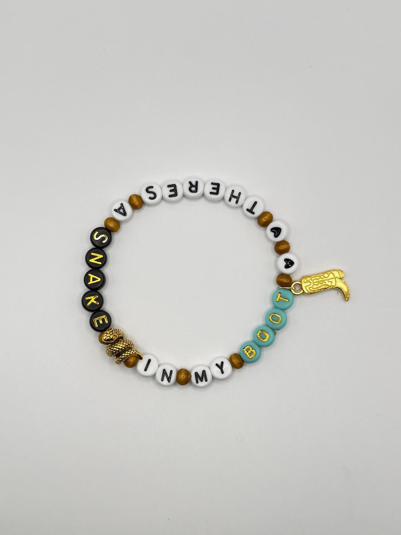 A beaded bracelet featuring white, black, and turquoise letter beads spelling out THERE'S A SNAKE IN MY BOOT. The design includes brown spacer beads, a gold-toned snake charm, and a gold-toned cowboy boot charm attached to the side.