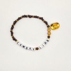 May include: A beaded bracelet featuring dark brown wooden beads, white letter beads spelling COWBOY LIKE, and tan letter beads spelling ME. The bracelet includes small gold-toned spacer beads and a gold-toned cowboy hat charm attached to the side.