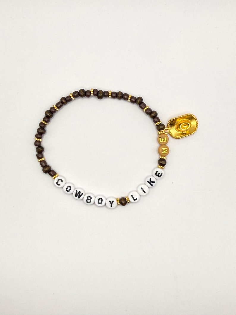 May include: A beaded bracelet featuring dark brown wooden beads, white letter beads spelling COWBOY LIKE, and tan letter beads spelling ME. The bracelet includes small gold-toned spacer beads and a gold-toned cowboy hat charm attached to the side.