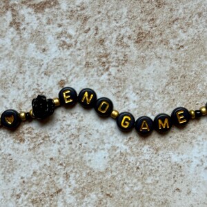 Pode incluir: Uma pulseira de contas pretas com detalhes dourados. A pulseira diz "END GAME" em letras douradas em contas circulares pretas. Uma pequena rosa preta tamb&eacute;m est&aacute; inclu&iacute;da no design.