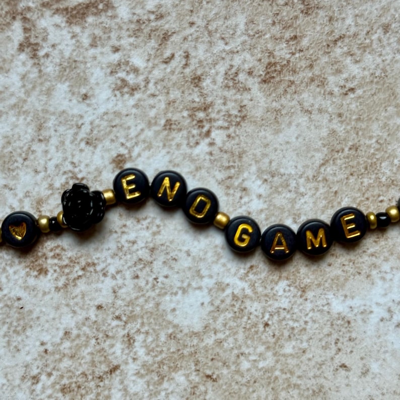Pode incluir: Uma pulseira de contas pretas com detalhes dourados. A pulseira diz "END GAME" em letras douradas em contas circulares pretas. Uma pequena rosa preta tamb&eacute;m est&aacute; inclu&iacute;da no design.