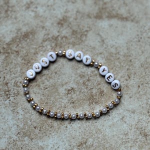 Pode incluir: Pulseira de contas brancas com as palavras "JUST SAY YES" escritas em letras douradas. A pulseira &eacute; feita de pequenas contas brancas e douradas, e &eacute; exibida em um fundo neutro.