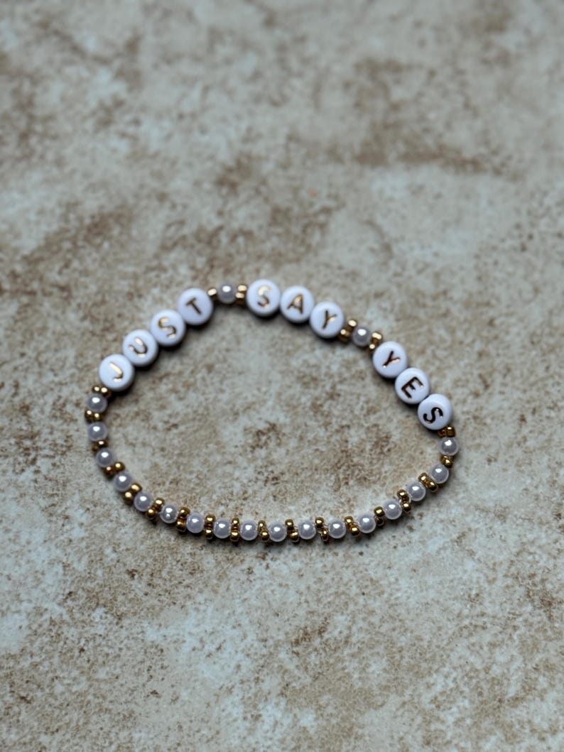 Pode incluir: Pulseira de contas brancas com as palavras "JUST SAY YES" escritas em letras douradas. A pulseira &eacute; feita de pequenas contas brancas e douradas, e &eacute; exibida em um fundo neutro.