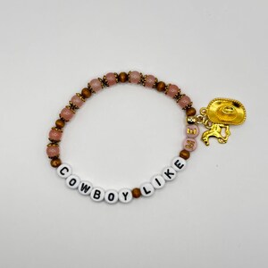 May include: A beaded bracelet featuring alternating pink and brown beads, white letter beads spelling COWBOY LIKE ME, and a gold-toned charm depicting a cowboy hat and a horse. The bracelet is circular and constructed with a flexible elastic cord.