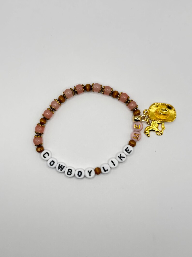 May include: A beaded bracelet featuring alternating pink and brown beads, white letter beads spelling COWBOY LIKE ME, and a gold-toned charm depicting a cowboy hat and a horse. The bracelet is circular and constructed with a flexible elastic cord.