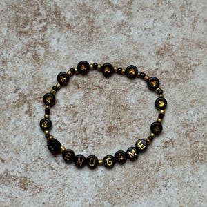 Pode incluir: Uma pulseira de contas pretas com detalhes dourados e a palavra "ENDGAME" escrita em letras douradas. A pulseira tamb&eacute;m apresenta contas pretas com formas de cora&ccedil;&atilde;o douradas.