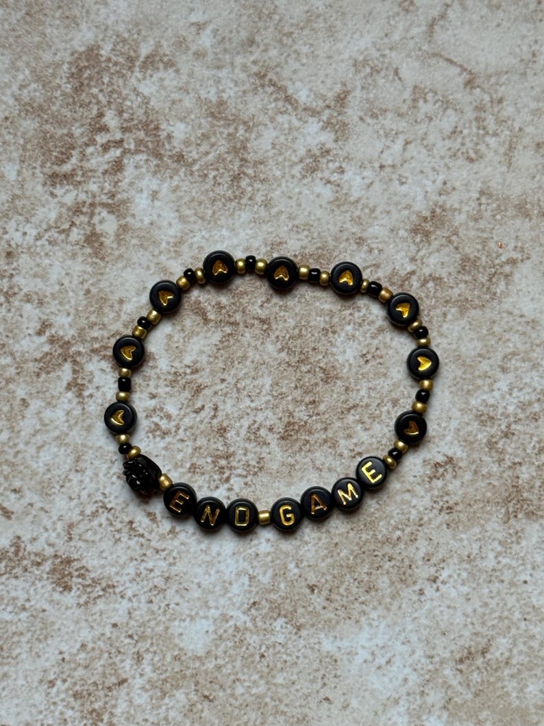 Pode incluir: Uma pulseira de contas pretas com detalhes dourados e a palavra "ENDGAME" escrita em letras douradas. A pulseira tamb&eacute;m apresenta contas pretas com formas de cora&ccedil;&atilde;o douradas.