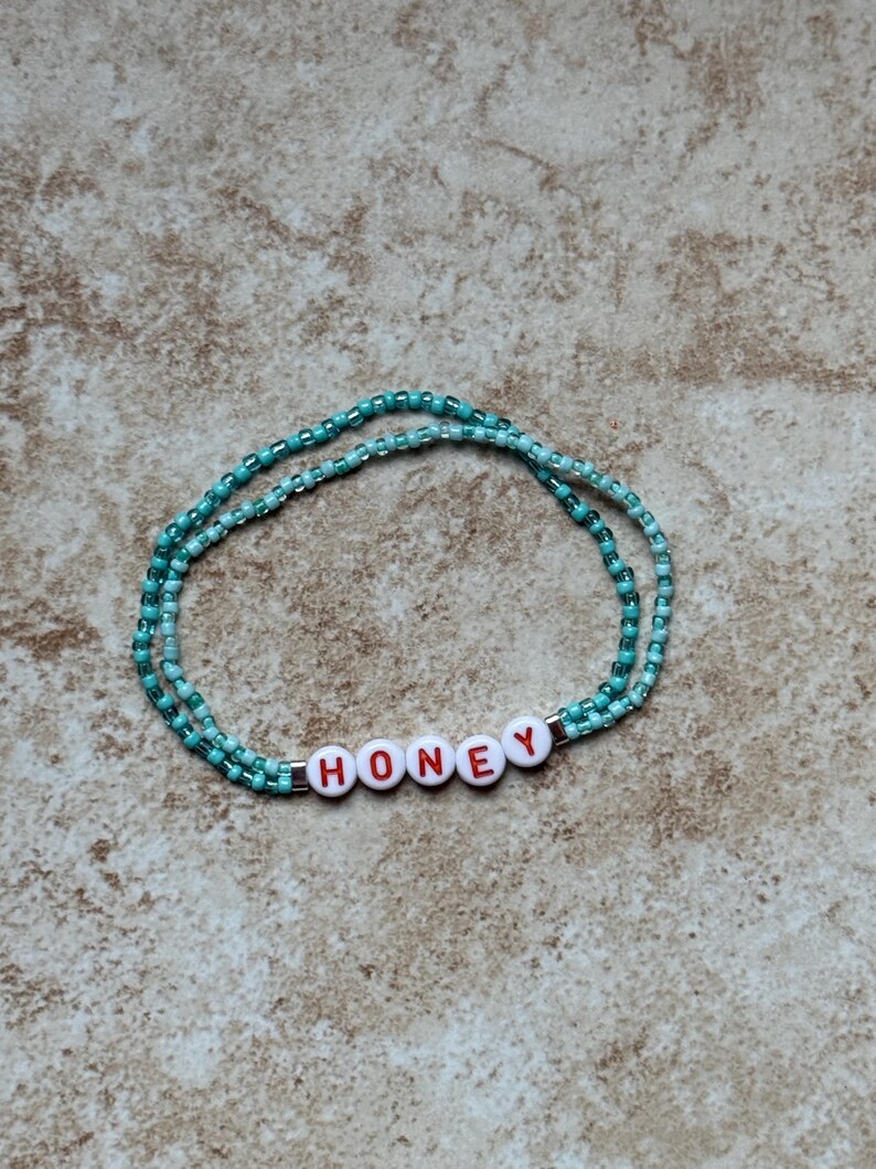 Pode incluir: Uma pulseira de contas turquesa com a palavra "HONEY" escrita em letras vermelhas em contas brancas. A pulseira &eacute; feita de pequenas contas e &eacute; exibida em uma superf&iacute;cie de tom neutro.