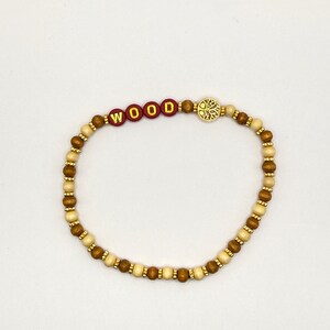 May include: A beaded bracelet featuring alternating light and dark brown wooden beads, small gold-toned spacers, four red circular beads with the gold lettered text WOOD, and a circular gold-toned charm depicting a tree with intricate branches.