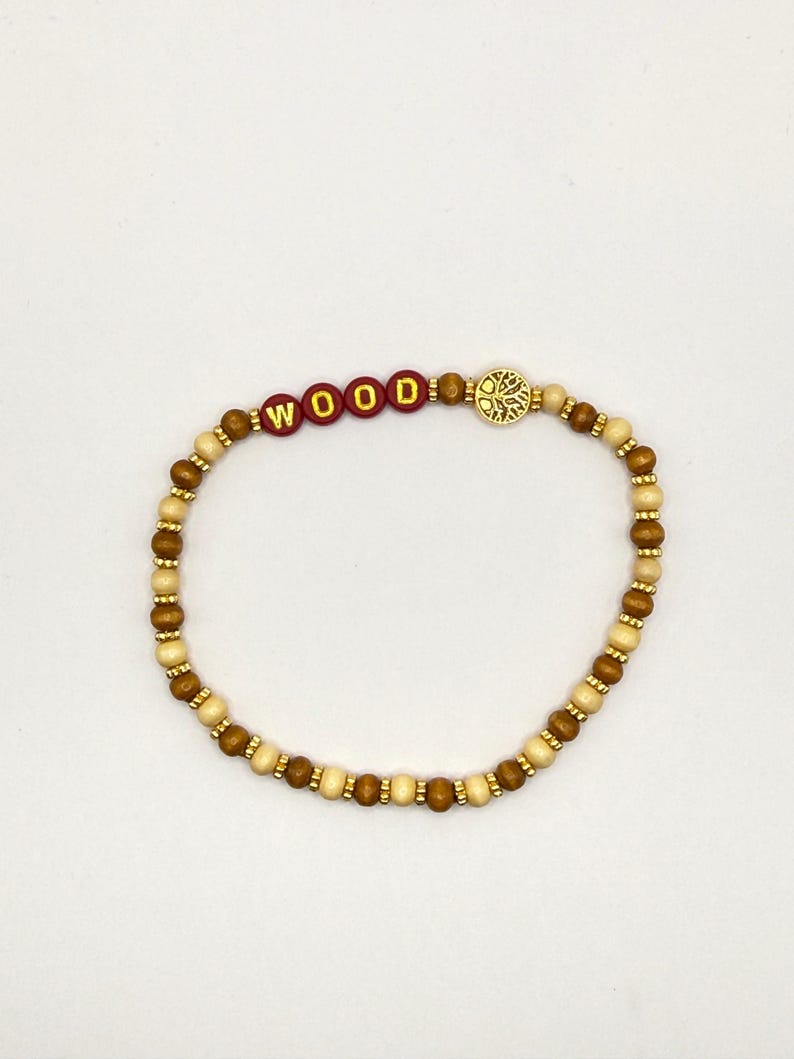 May include: A beaded bracelet featuring alternating light and dark brown wooden beads, small gold-toned spacers, four red circular beads with the gold lettered text WOOD, and a circular gold-toned charm depicting a tree with intricate branches.
