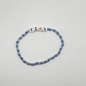 May include: A beaded bracelet featuring a repeating pattern of small, translucent blue and silver glass beads. The center of the bracelet includes four white round beads with black lettering and a gold star, spelling out the text TS star 5.
