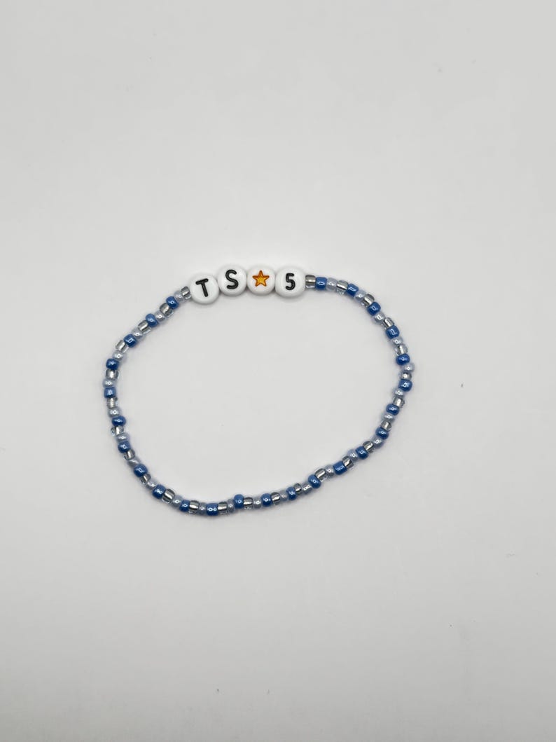 May include: A beaded bracelet featuring a repeating pattern of small, translucent blue and silver glass beads. The center of the bracelet includes four white round beads with black lettering and a gold star, spelling out the text TS star 5.