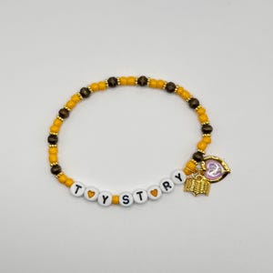 May include: A beaded bracelet featuring alternating yellow and dark brown round beads, with white letter beads spelling TOY STORY separated by small yellow heart beads. A gold-toned book charm and a heart-shaped charm hang from the bracelet.