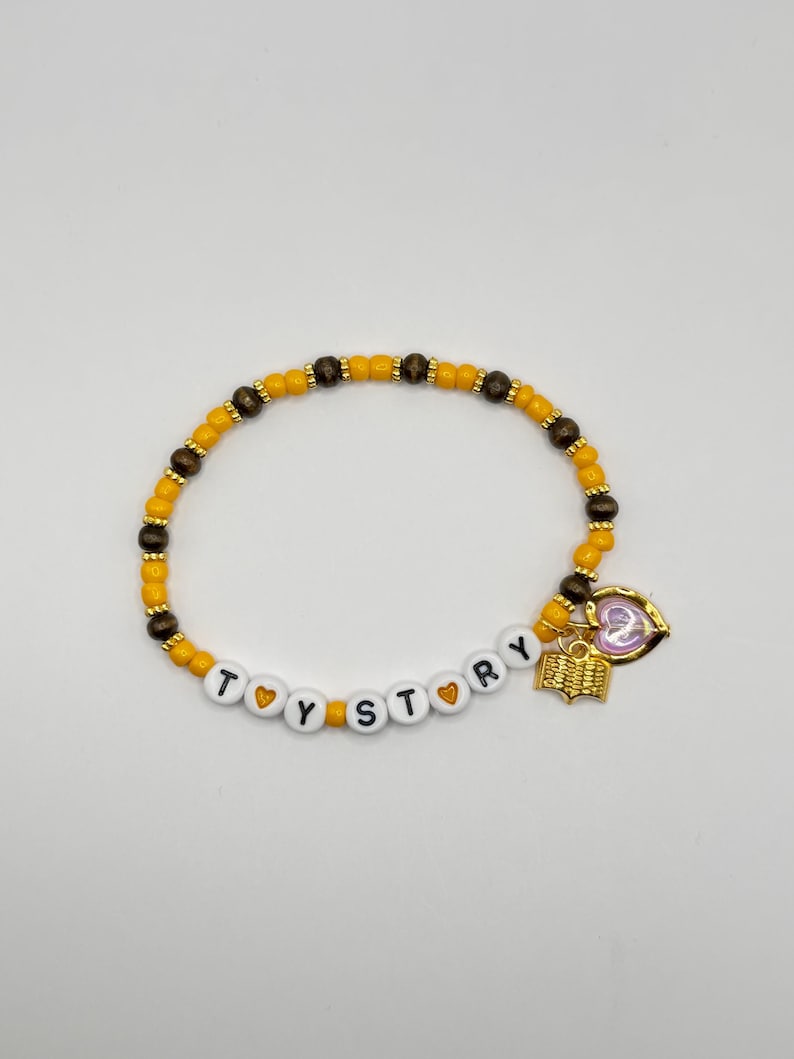 May include: A beaded bracelet featuring alternating yellow and dark brown round beads, with white letter beads spelling TOY STORY separated by small yellow heart beads. A gold-toned book charm and a heart-shaped charm hang from the bracelet.