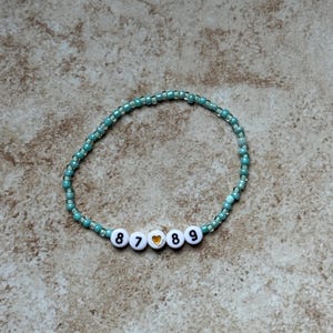 Pode incluir: Uma pulseira de contas turquesa e transparentes. A pulseira apresenta contas de letras brancas que soletram os n&uacute;meros "8789" com uma conta de cora&ccedil;&atilde;o dourado. A pulseira &eacute; exibida em uma superf&iacute;cie de tom neutro.
